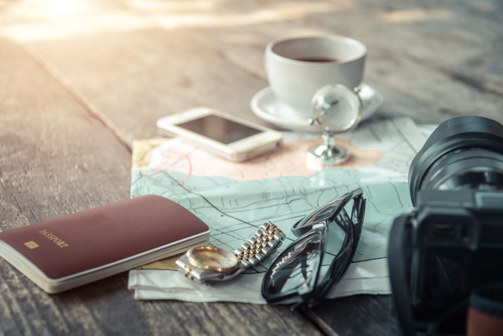 Photograph of travel essentials: a phone, map, passport, camera, sunglasses, watch, and a cup of coffee.